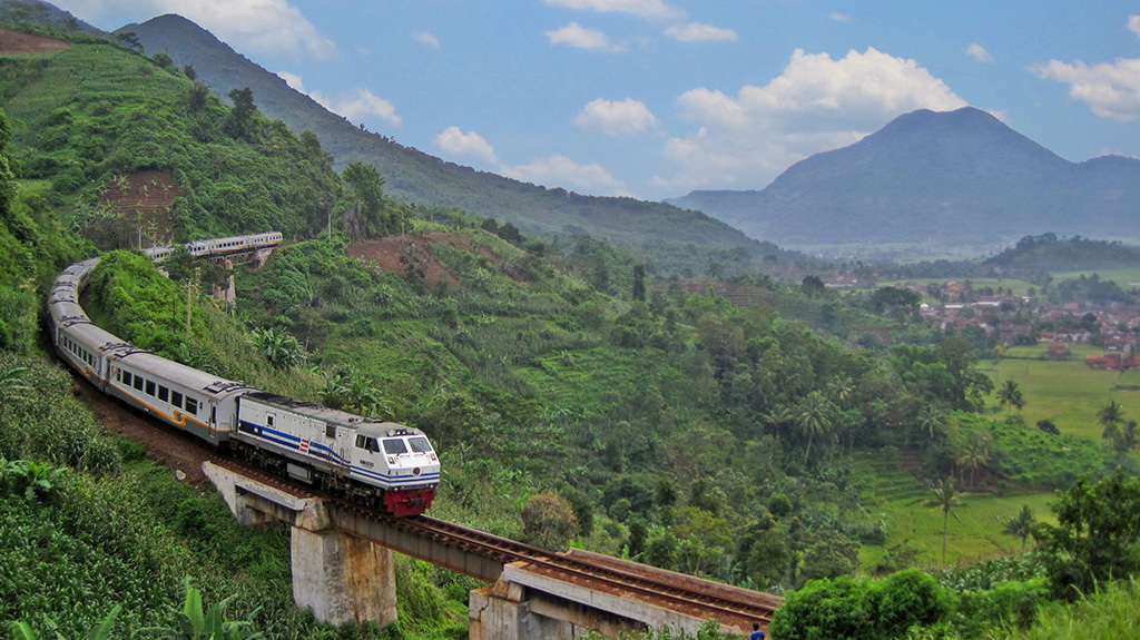 Scenic train journey through lush green hills and valleys with mountains in the background.