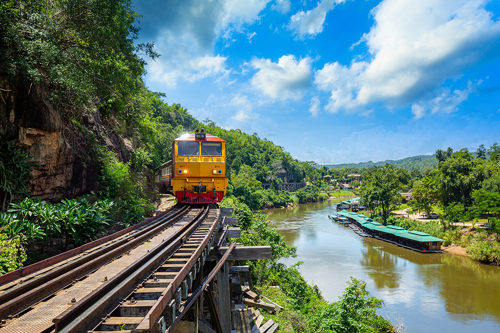 Train on the historic Death Railway bridge over the River Kwai in Thailand, surrounded by lush greenery and scenic river views.