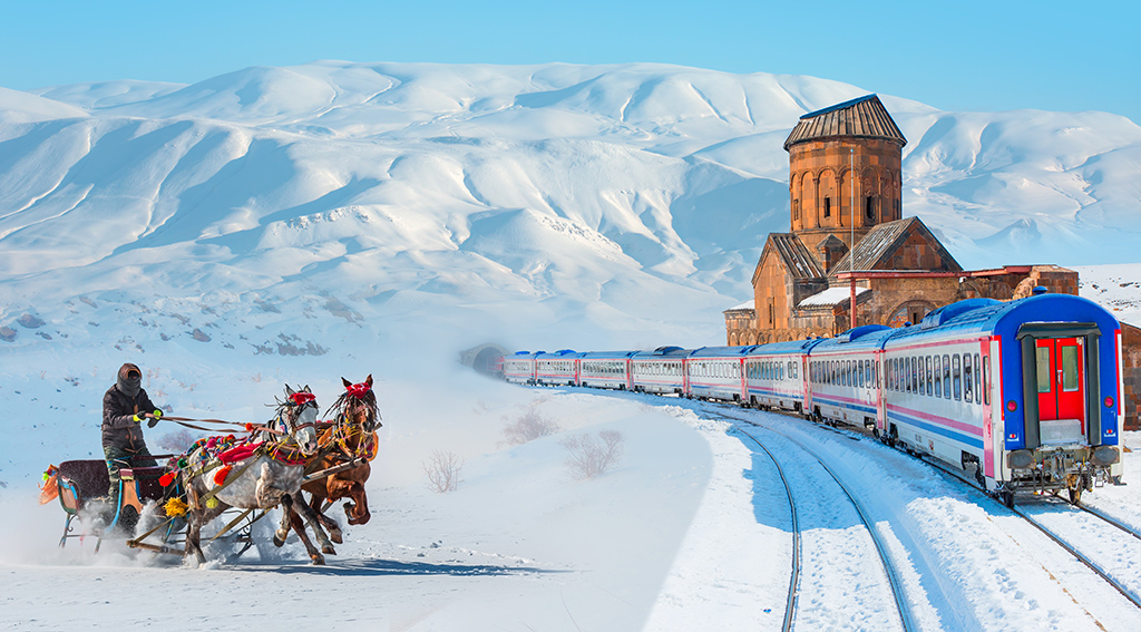 A scenic winter landscape featuring a horse-drawn sleigh and a modern train passing by an ancient church with snow-covered mountains in the background.