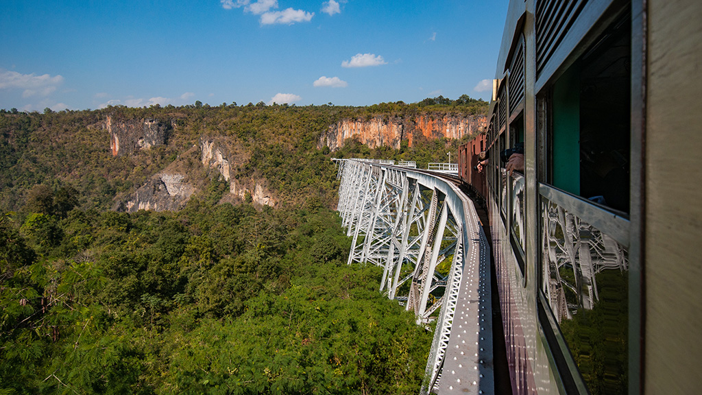 Scenic train journey over Goteik Viaduct in Myanmar with lush green forest and rocky cliffs in the background.