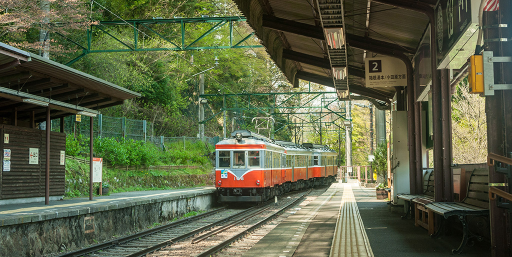 Vintage red and white train arriving at a quaint, scenic railway station surrounded by lush greenery and traditional architecture.