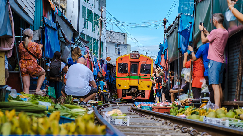 A vibrant train moves through the bustling Maeklong Railway Market in Thailand, with vendors and tourists closely lining the tracks.