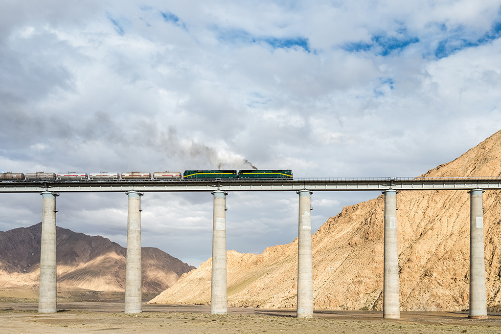 Freight train crossing a high elevated bridge in a mountainous desert landscape under a cloudy sky.