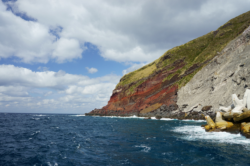 Coastal cliff with red and green layers, blue ocean waves, and concrete tetrapods under a partly cloudy sky.